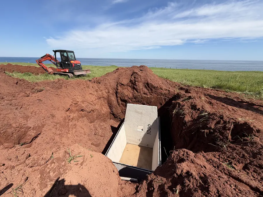 Mini excavator above an open red-soil trench beside the sea